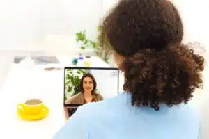 Frau mit braunen Locken im Zopf und blauer Bluse von hinten sitzt an weißem Schreibtisch mit gelber Kaffeetasse und Laptop. Im Laptop-Bildschirm eine sprechende Frau mit braunem Haar, braunem Cardigan und rotem Top, hinter ihr Zimmerpflanze.