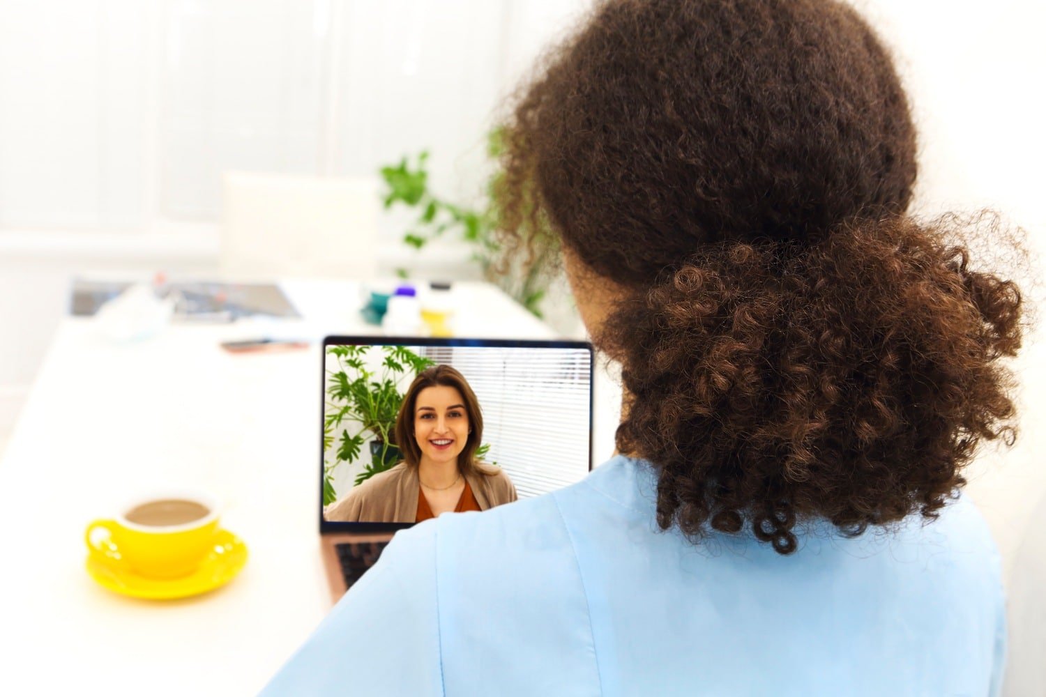 Frau mit braunen Locken im Zopf und blauer Bluse von hinten sitzt an weißem Schreibtisch mit gelber Kaffeetasse und Laptop. Im Laptop-Bildschirm eine sprechende Frau mit braunem Haar, braunem Cardigan und rotem Top, hinter ihr Zimmerpflanze.