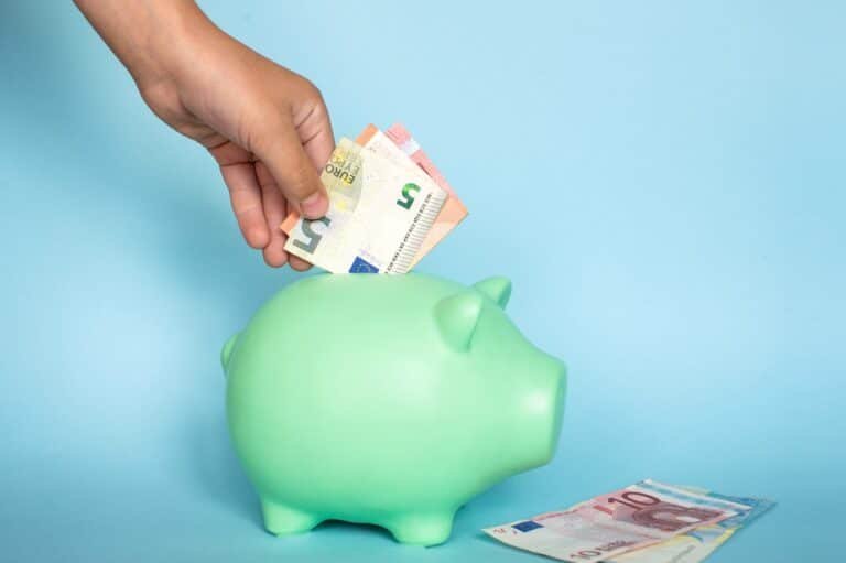 Hand legt einen Fünf- und einen Zehn-Euro-Schein in ein grünes Sparschwein. Blauer Hintergrund.