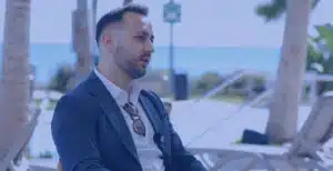 Mann im dunkelblauen Anzug mit weißem Hemd sitzt in einem Interview an einer Strandpromenade mit Palmen und Meer im Hintergrund.